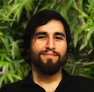 Man with dark hair and beard wearing a black shirt, photographed outdoors with green foliage background