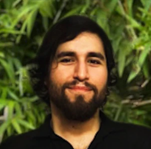 Man with dark hair and beard wearing a black shirt, looking at camera with green foliage background