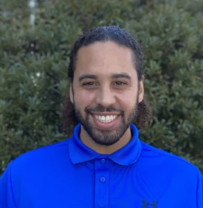 Man with dark hair and beard wearing a bright blue polo shirt, smiling at camera against a blurred green background.