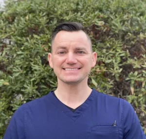 Man with dark hair smiling at camera wearing dark blue v-neck shirt against blurred green foliage