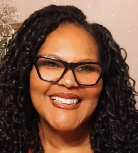 Woman with dark curly hair and glasses wearing dark clothing, smiling at camera against a blurred background.
