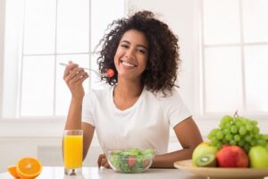 young woman seated at kitchen table with fresh healthy fruits and vegetables for her meal