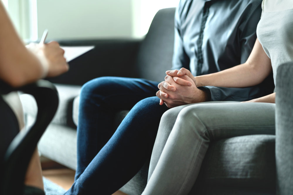 close up of a couple's joined hands as they sit on couch while talking to a therapist