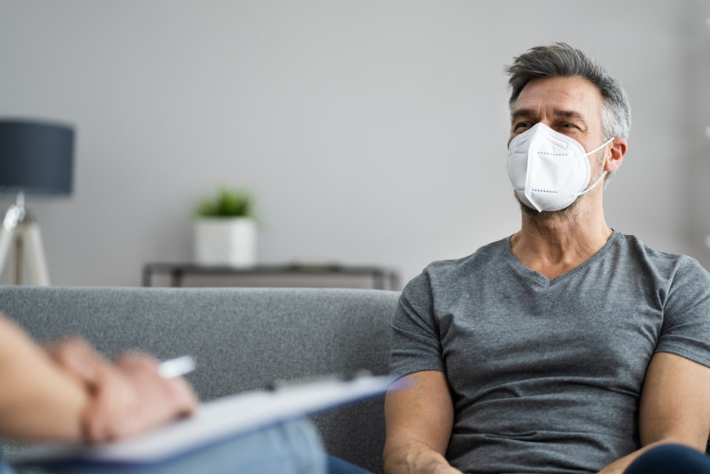 older adult male wearing n-95 mask while seated on couch talking to therapist