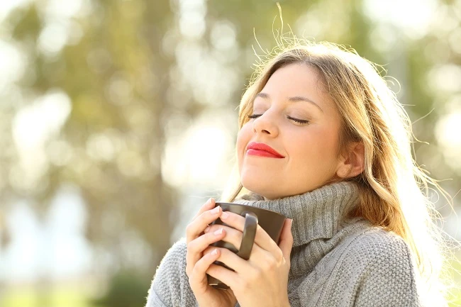 Close-up portrait of a woman with reddish-blonde hair in sunlight holding a warm mug with eyes closed and pink lipstick