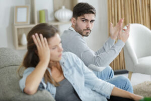 man and woman sitting on couch but clearly annoyed with each other as the man is throwing both hands up in the air and the woman has turned away and has a look of disdain