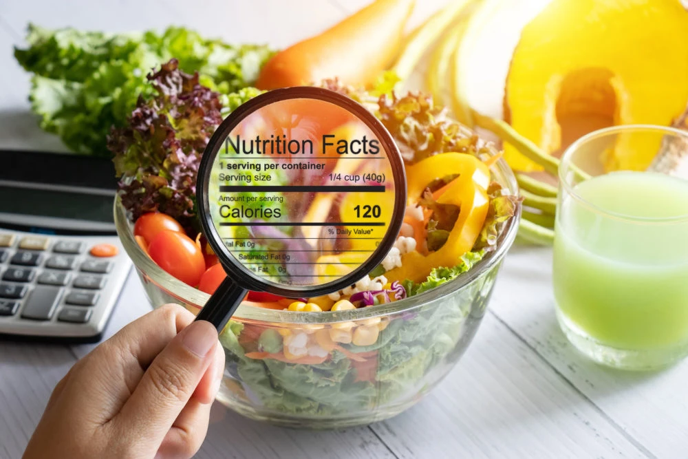 Hands holding magnifying glass over bowl of salad with nutrition facts label, laptop and juice glass visible