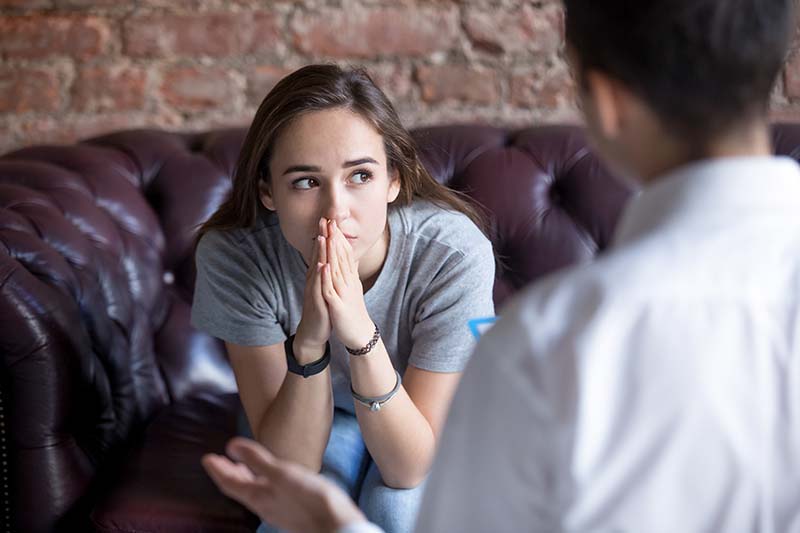 young girl sitting on couch with hands pressed against mouth thoughtfully while therapist counsels her