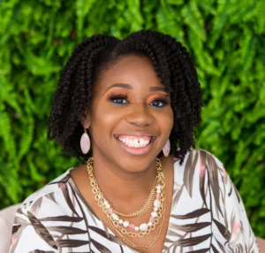 Woman with dark curly hair wearing a striped shirt and pearl necklace, smiling at camera against a green background.