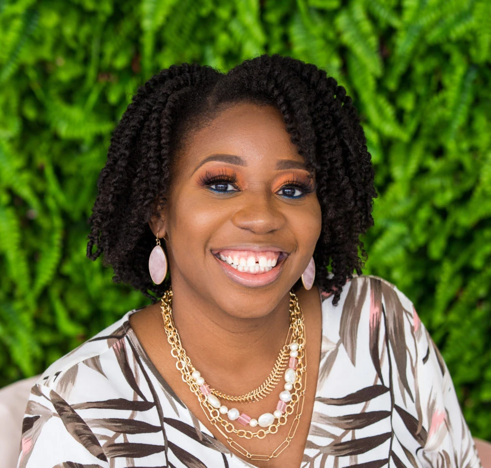 Woman with dark curly hair wearing a striped shirt and pearl necklace, smiling at camera against a green background.