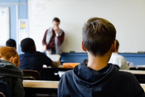 view of the back of a male child's head as he sits in a classroom with teacher out of focus at front of room