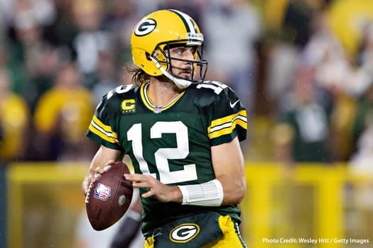 Football player wearing green and yellow uniform and helmet, holding a football, standing on field with blurred crowd in background
