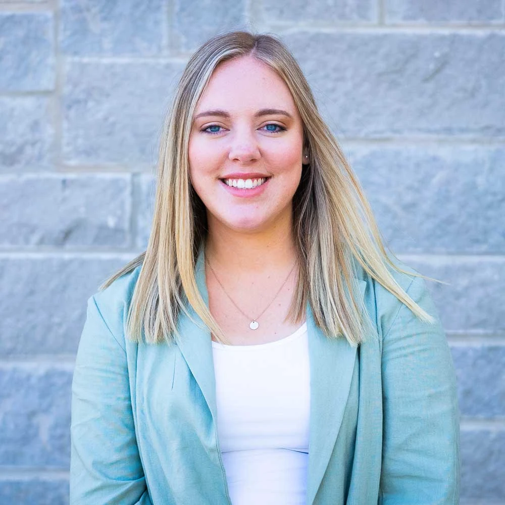 Woman with long blonde and light brown hair wearing a light green cardigan over white top, smiling at camera against blue tile background