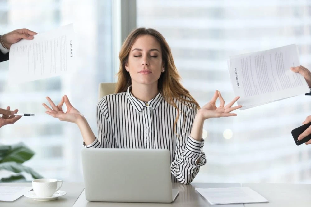 Woman with reddish-brown hair at laptop surrounded by papers being held by other hands, expressing stress and overwhelm