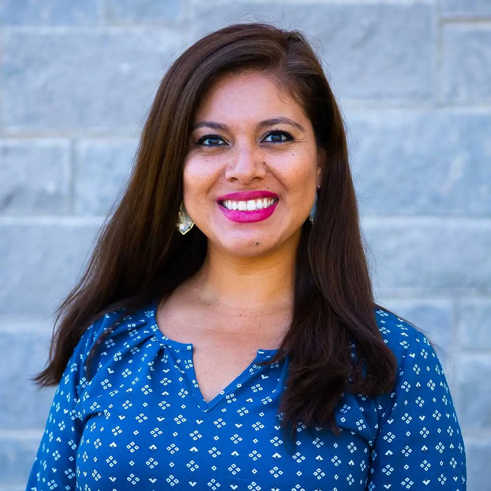 Woman with long dark hair wearing a blue patterned top, smiling at camera against a blue brick wall background