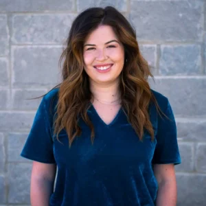 Woman with wavy dark brown hair wearing a navy blue v-neck shirt, smiling at camera against brick wall