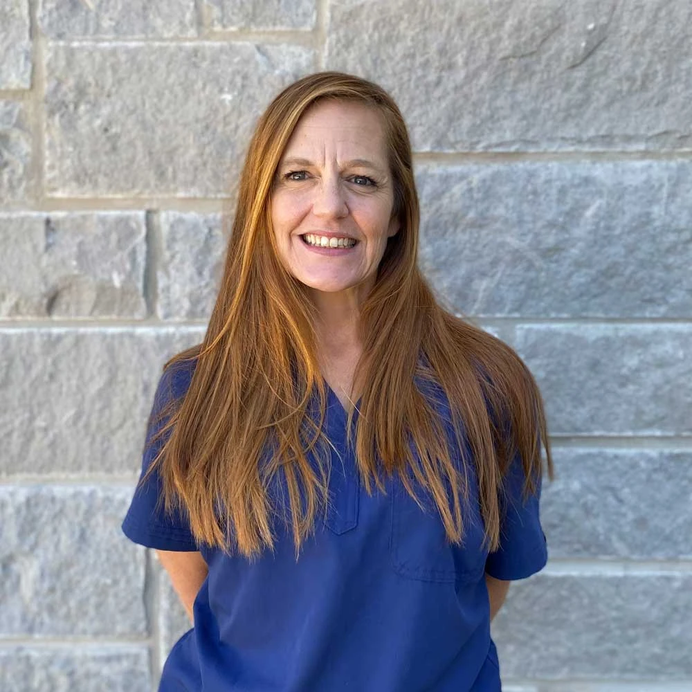 Smiling woman with long reddish-brown hair wearing a dark blue scrubs top against a brick wall background