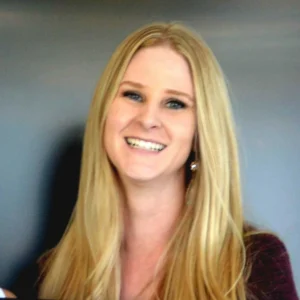Woman with long blonde hair wearing a dark burgundy top, smiling at camera against a neutral gray background