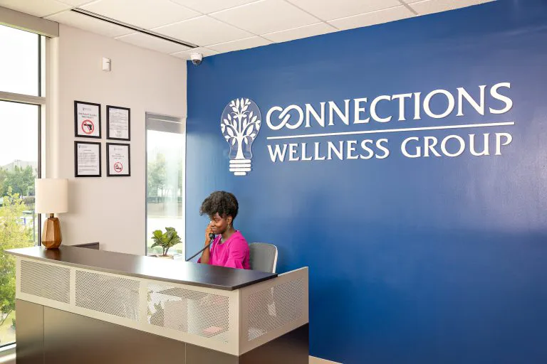 Professional reception desk with woman in bright pink top sitting at counter, blue Connections Wellness Group logo wall