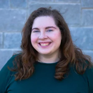 Woman with long brown wavy hair smiling at camera wearing dark green shirt against brick wall background