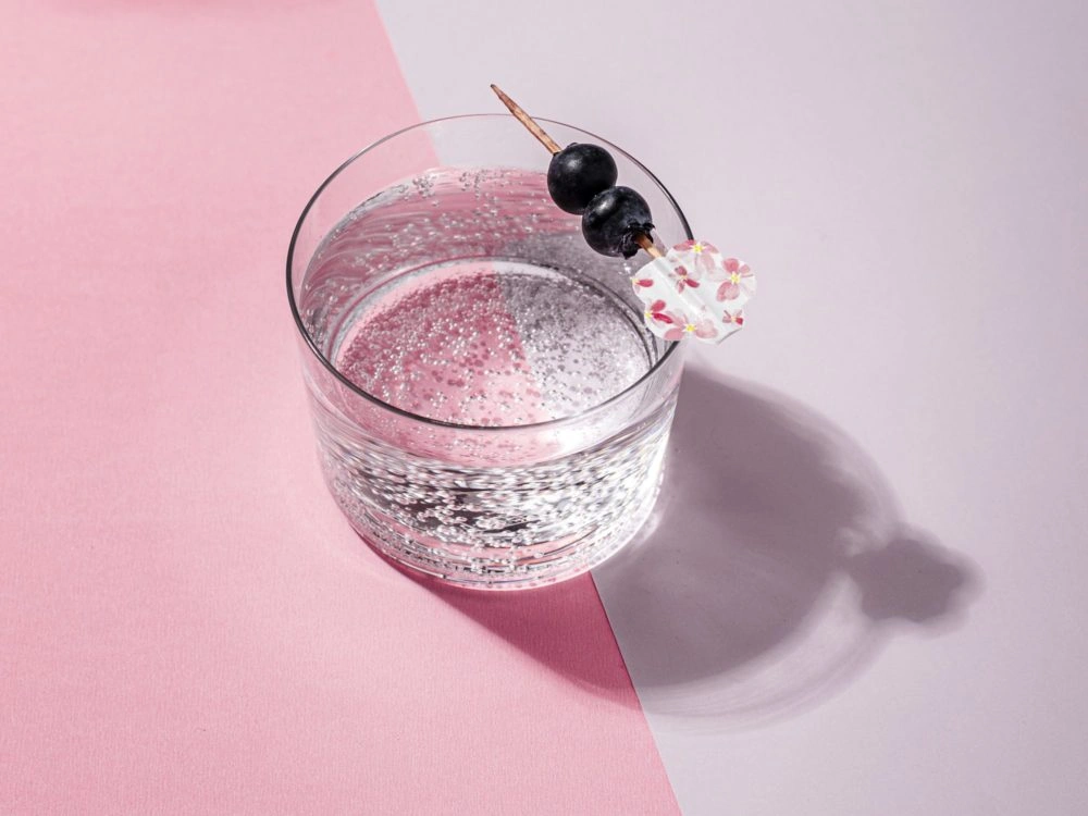 Clear cocktail with pink and silver glitter on pink and white background with black berries and flower garnish