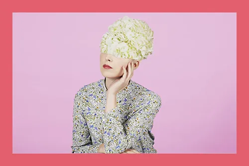 Portrait of person with white hydrangea flowers covering head, wearing patterned jacket against pink background