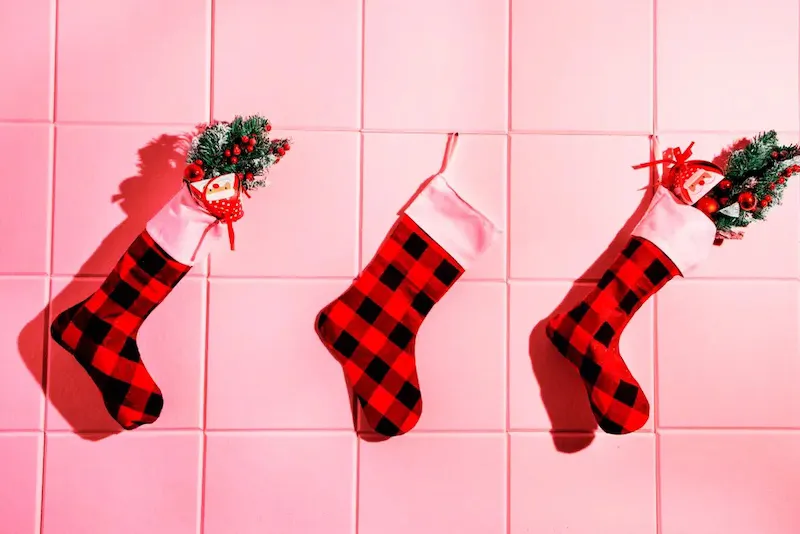 Three red and black plaid Christmas stockings with greenery and bows hanging against bright pink tile wall
