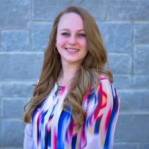 Woman with long reddish-brown hair wearing colorful striped blouse smiling against brick wall