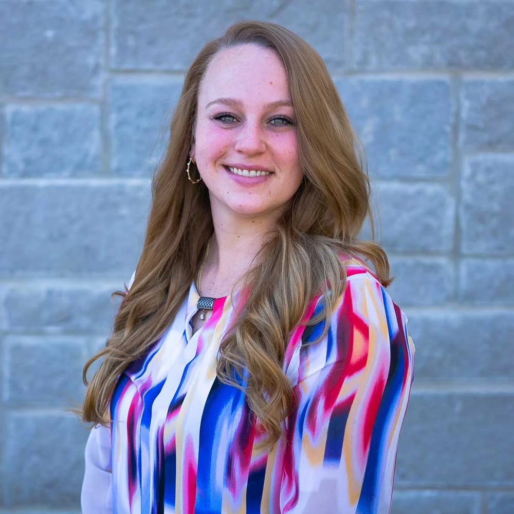 Woman with long reddish-brown hair wearing colorful striped blouse smiling against brick wall