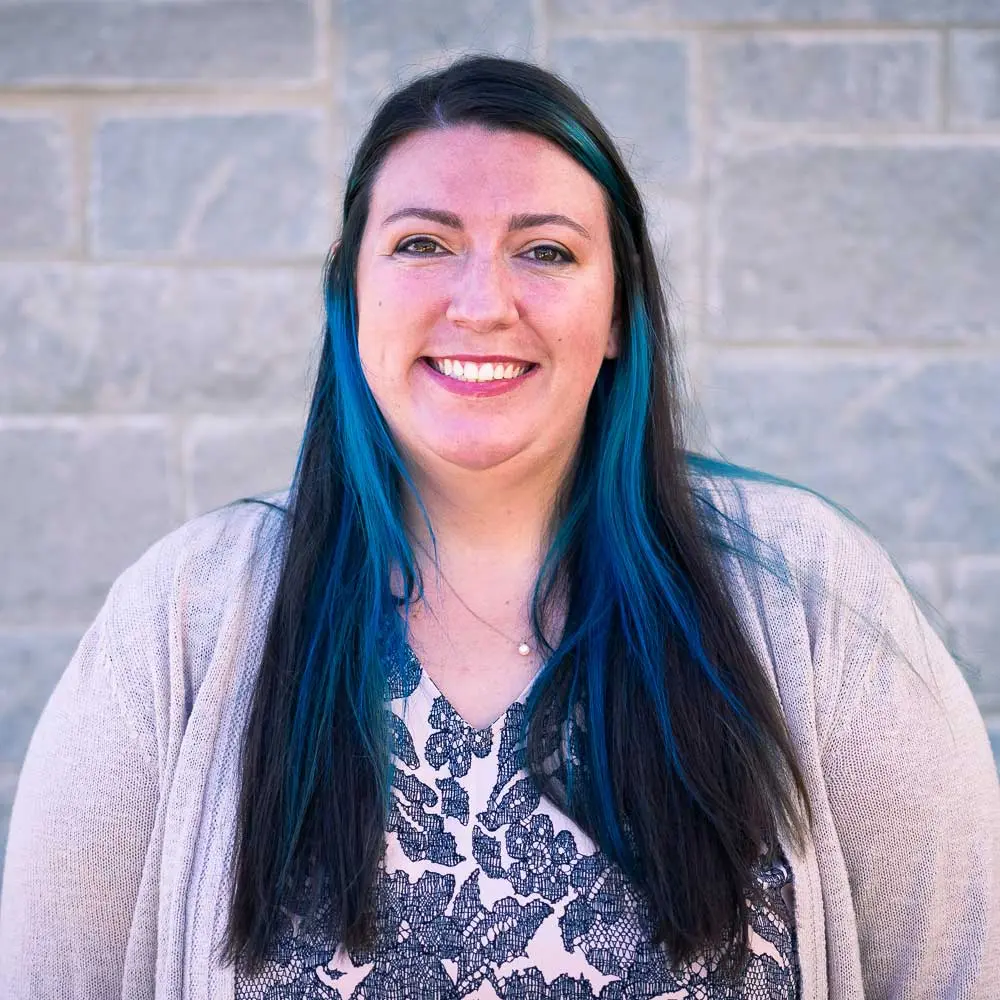 Woman with long dark hair with blue highlights wearing a beige cardigan over patterned top, smiling in front of brick wall