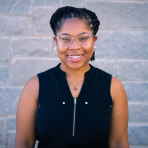 Woman with dark braided hair wearing glasses and nose ring, smiling at camera against brick wall background