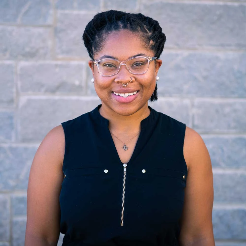 Woman with dark braided hair wearing glasses and nose ring, smiling at camera against brick wall background