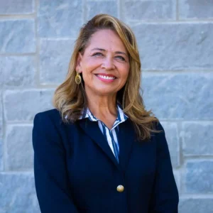 Mature woman with wavy reddish-brown hair wearing dark blazer and blue striped blouse smiling at camera against gray stone wall