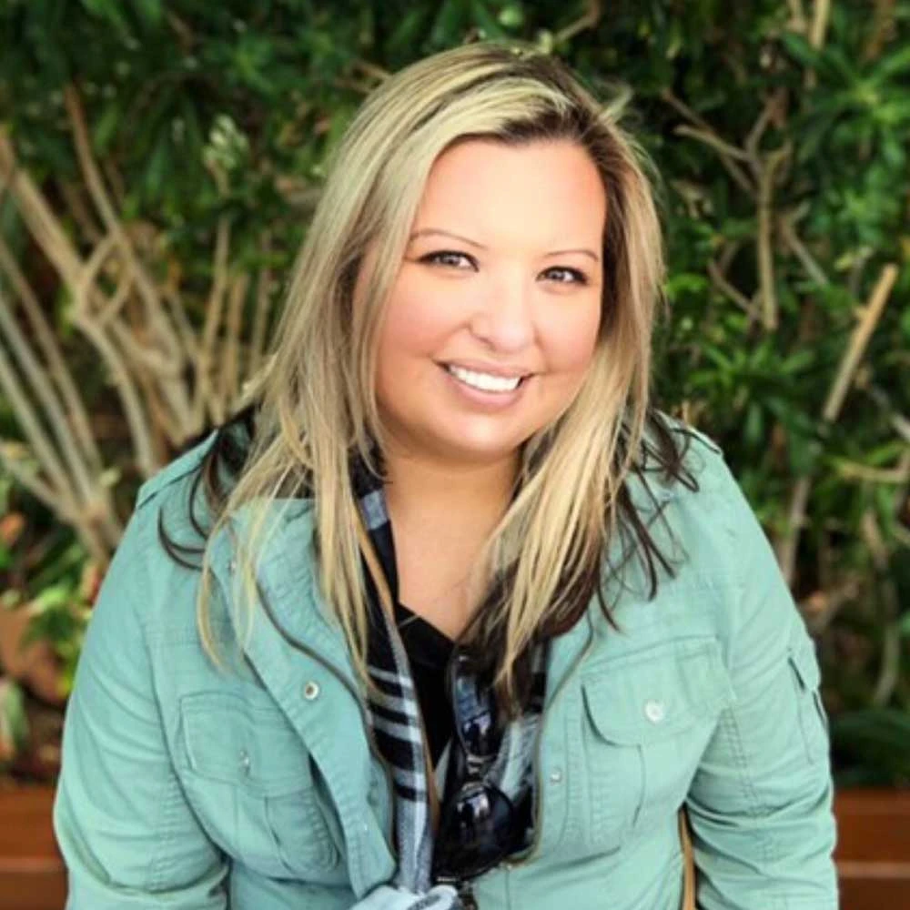 Woman with blonde hair wearing a light green button-up shirt over a dark striped top, smiling at camera with green foliage background