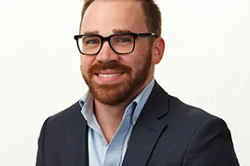 Portrait of man with glasses wearing dark blazer and light blue shirt, smiling at camera against white background