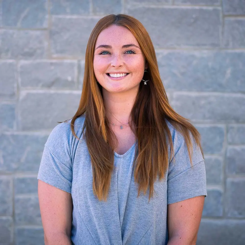 Woman with long reddish-brown hair smiling at camera, wearing light blue t-shirt, gray brick wall background