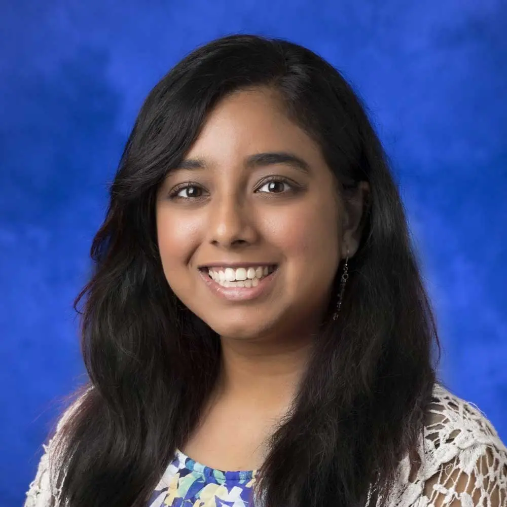 Woman with long dark hair smiling at camera wearing patterned white and blue top against blue background