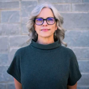 Woman with shoulder-length gray and dark hair wearing glasses and dark turtleneck, smiling at camera against brick wall
