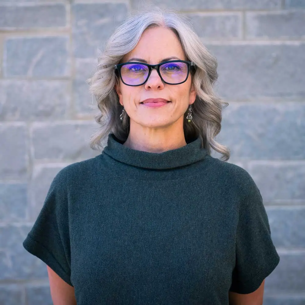 Woman with shoulder-length gray and dark hair wearing glasses and dark turtleneck, smiling at camera against brick wall