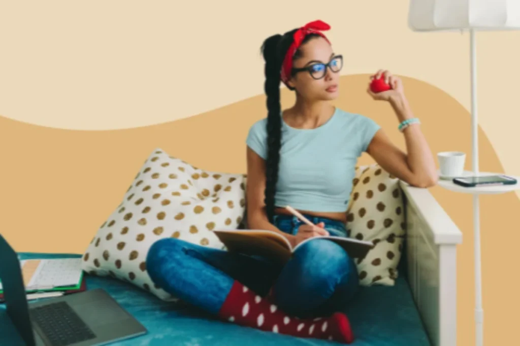Woman wearing light blue shirt and red headband sitting on patterned couch holding red object with laptop nearby