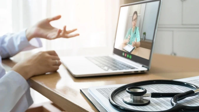 Doctor's hands gesturing during virtual video consultation on laptop screen with stethoscope visible on desk