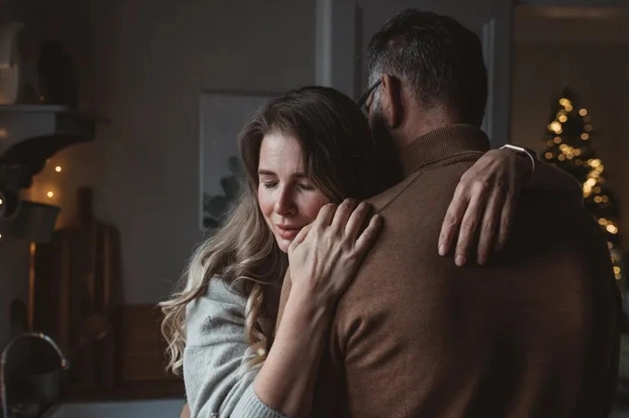 Man in brown turtleneck hugging woman from behind in indoor setting with blurred holiday lights in background
