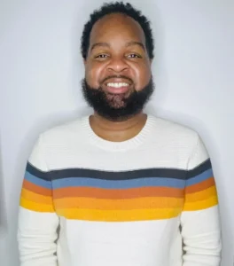 Smiling man with dark beard wearing white and rainbow striped sweater in indoor portrait setting with white background.