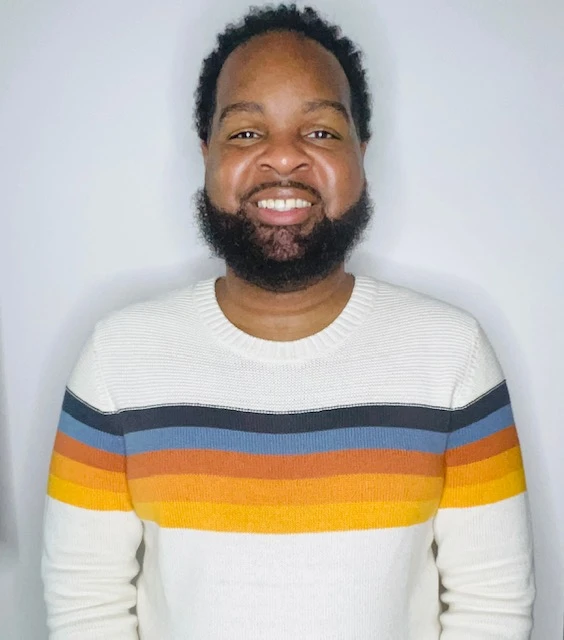 Smiling man with dark beard wearing white and rainbow striped sweater in indoor portrait setting with white background.