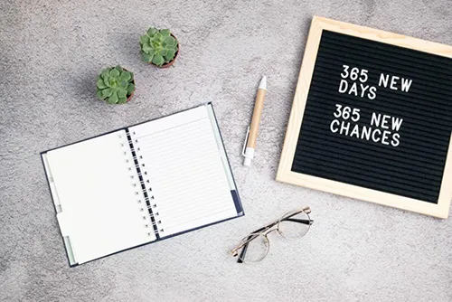 Flat lay workspace with open spiral notebook, two small succulents, wooden pen, eyeglasses, and letterboard displaying '365 new days 365 new chances...