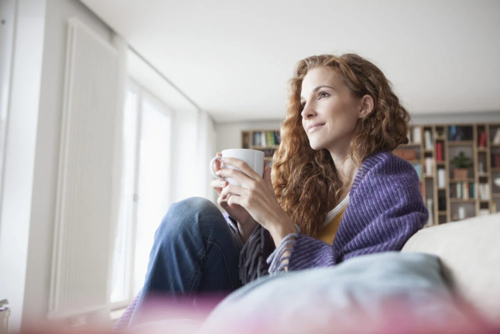 Woman with curly reddish-brown hair wearing purple cardigan holding white mug looking out window with bookshelves visible in soft natural light
