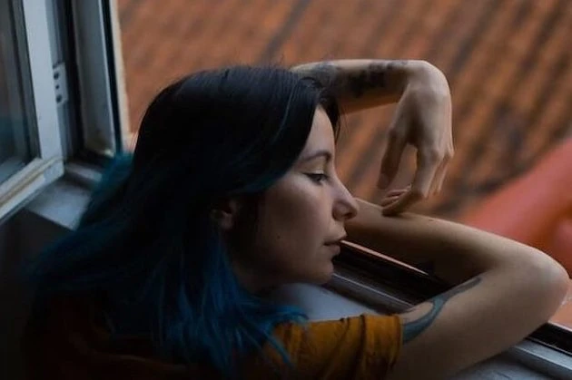 Woman with dark blue-tinted hair looking out window, resting head on hand, contemplative expression, warm lighting