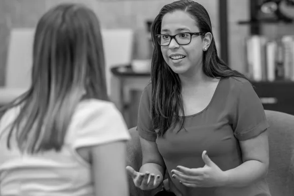 Black and white photograph of two women in conversation, one wearing glasses and brown shirt gesturing while speaking to woman with long hair facin...
