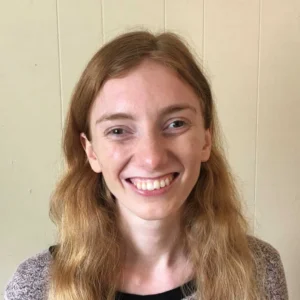 Woman with reddish-brown hair wearing a gray cardigan over black shirt, smiling at camera against plain beige wall