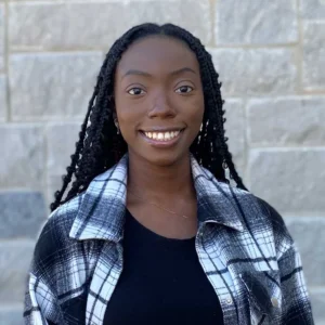 Professional headshot of a woman with long braided black hair smiling at camera wearing a black and white plaid shirt against a brick wall.